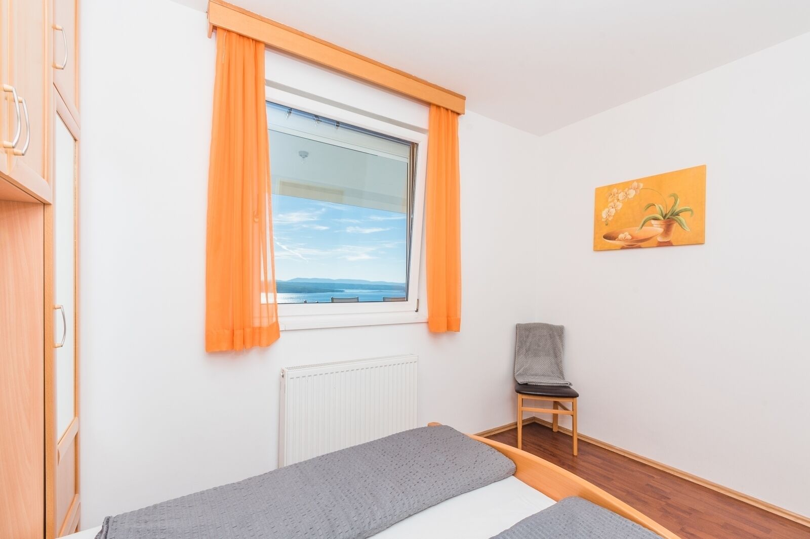 A room with two beds, a chair, a radiator, a window with orange curtains, and a painting on the wall.