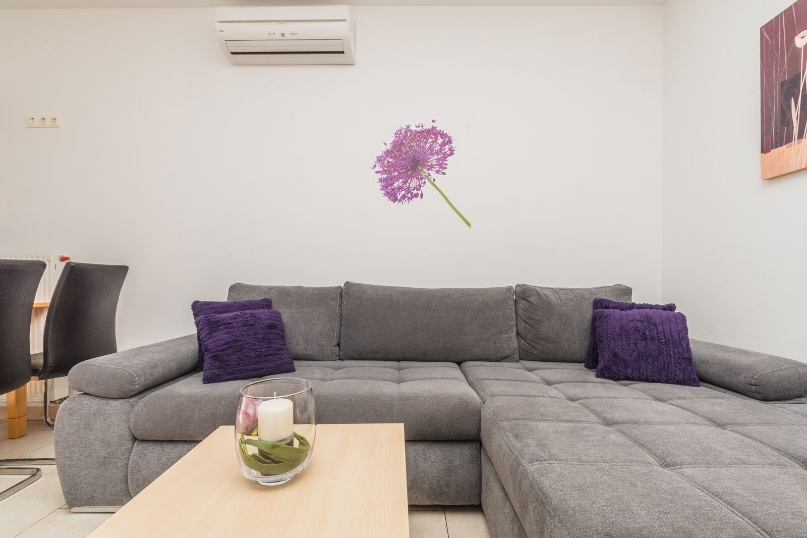 Una sala de estar moderna con un sofá seccional gris, dos almohadones morados, un candelabro de vidrio con vela y un aire acondicionado instalado en la pared.