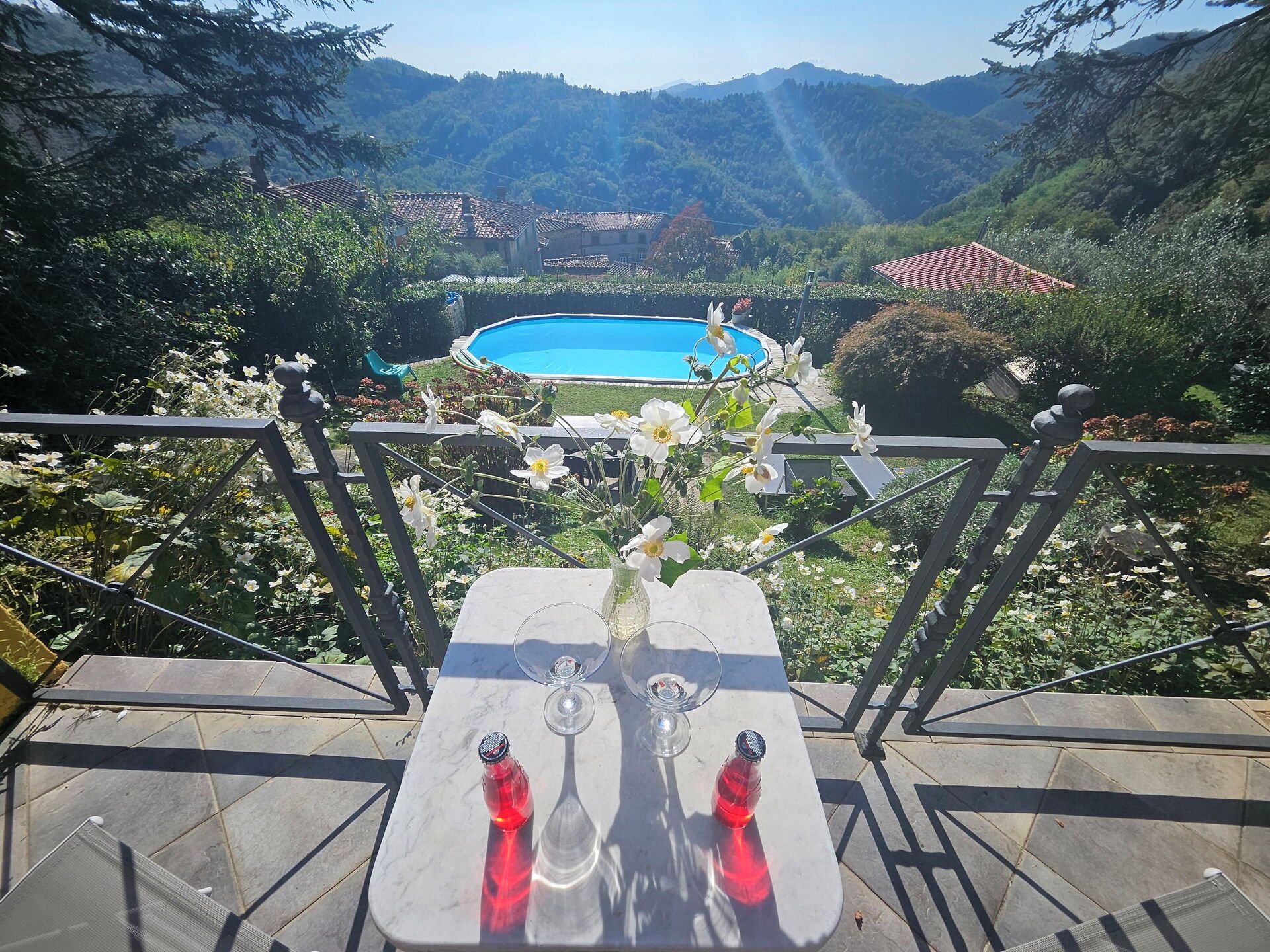 A terrace with a table, two glasses, and a vase with flowers. In the background, there is a swimming pool surrounded by greenery and houses.