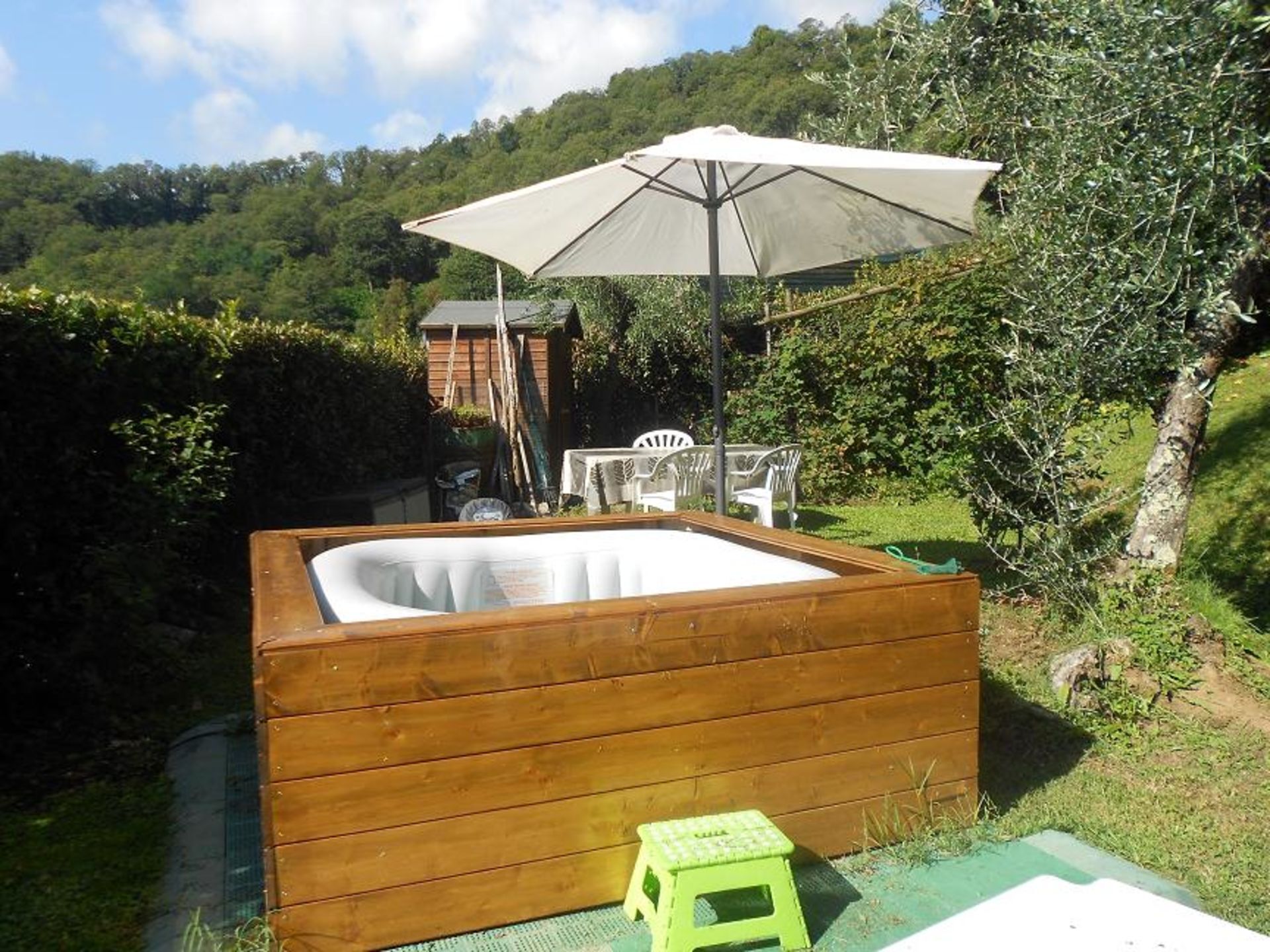 A wooden hot tub with a large umbrella, surrounded by greenery and a wooden fence.
