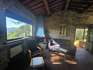 Un intérieur rustique en pierre, avec un bureau, un fauteuil rembourré, un canapé et une vue sur un paysage boisé par la fenêtre.