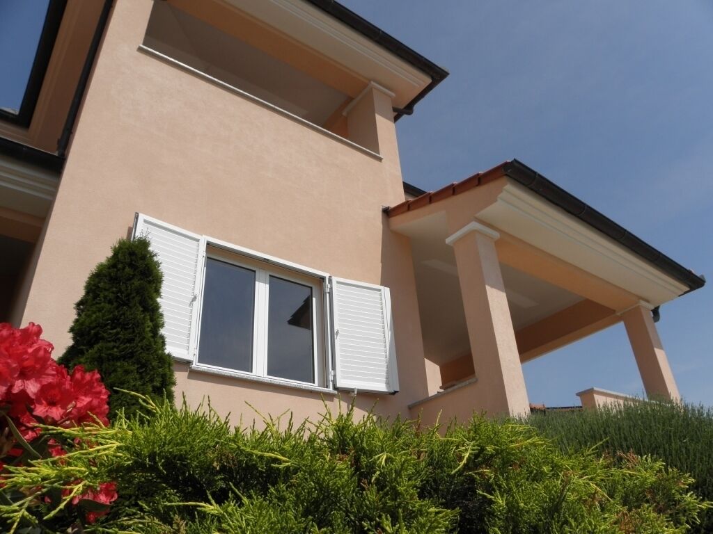 A two-story building with a balcony, a window with shutters, and a well-maintained garden with shrubs and flowers.
