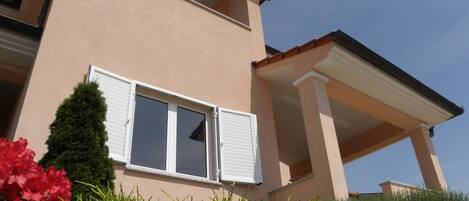 A two-story building with a balcony, a window with shutters, and a well-maintained garden with shrubs and flowers.