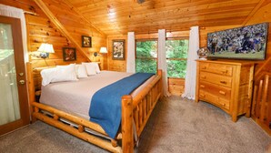 Un dormitorio de cabaña de madera con una cama, un aparador, un ventilador de techo y un televisor que muestra un partido de deportes.