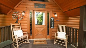 Entrada de una cabaña de madera con una puerta en forma de cruz, dos mecedoras con cojines blancos, un letrero de bienvenida y un tapete que dice '¡Hooray! ¡Estás aquí'.