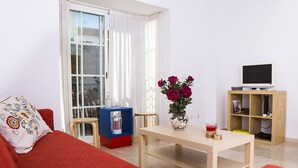 Ett vardagsrum med en röd soffa, ett soffbord i trä, en tv på ett tv-bord, en bokhylla och ett fönster med gardiner.