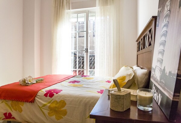A bedroom with a bed, a window with curtains, a bedside table with a candle, and a framed picture on the wall.
