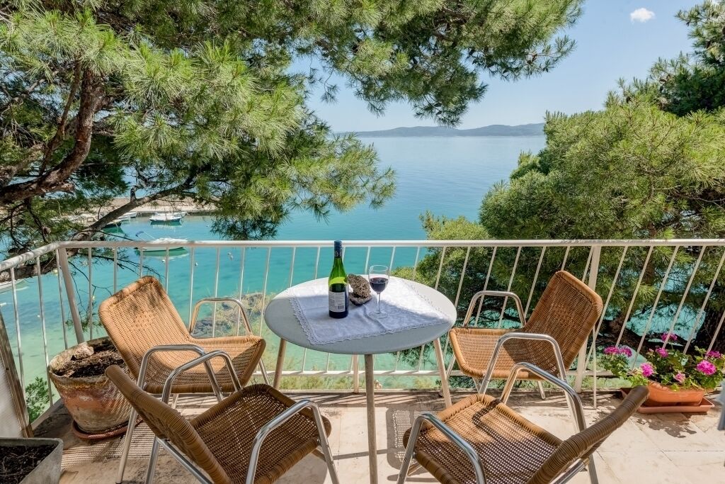 Un balcón con sillas de mimbre, una mesa con una botella y un vaso, y vistas al mar y a las montañas.