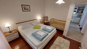 Un dormitorio con cama, mesitas de noche, una cuna, un tapete y una zona de cocina al fondo.