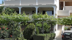 A two-story house with a balcony, a car parked in front, and a garden with grapevines and bushes.