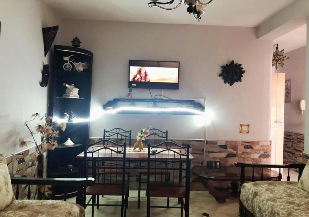 Een woonkamer met een aan de muur bevestigde televisie, een eettafel met stoelen, een bank en een decoratieve plank met beeldjes.