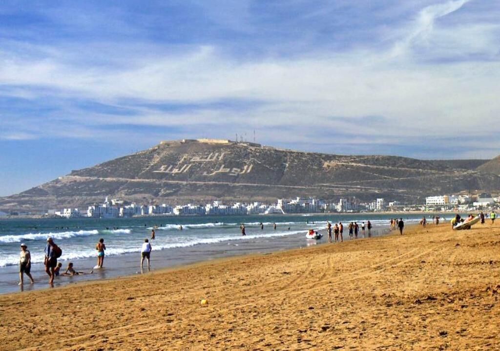 A beach with people walking and playing, a hill with Arabic script, and buildings in the background.