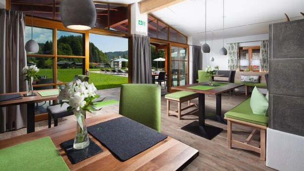 A modern hotel lobby with wooden flooring, green cushioned seating, and large windows offering a view of a green landscape. - Ferienwohnung  2 bis 2 Personen, 52 qm, Erdgeschoss, 1 separates Schlafzimmer, Terrasse (Schönau am Königssee)