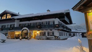 A large, multi-story building with a snow-covered exterior, balconies with railings, and a lit entrance. - Ferienwohnung  2 bis 2 Personen, 52 qm, Erdgeschoss, 1 separates Schlafzimmer, Terrasse (Schönau am Königssee)