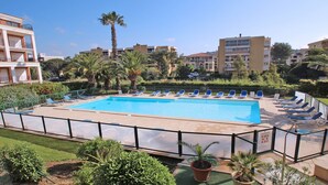 Ein rechteckiger Swimmingpool mit Fliesen umrandet, umgeben von einem Metallzaun. Entlang des Poolrands reihen sich Liegestühle.