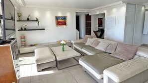 Ein modernes Wohnzimmer mit einer Couch, einem Couchtisch und einem Fernseher. Dekorative Pflanzen und ein Gemälde zieren die Wand.