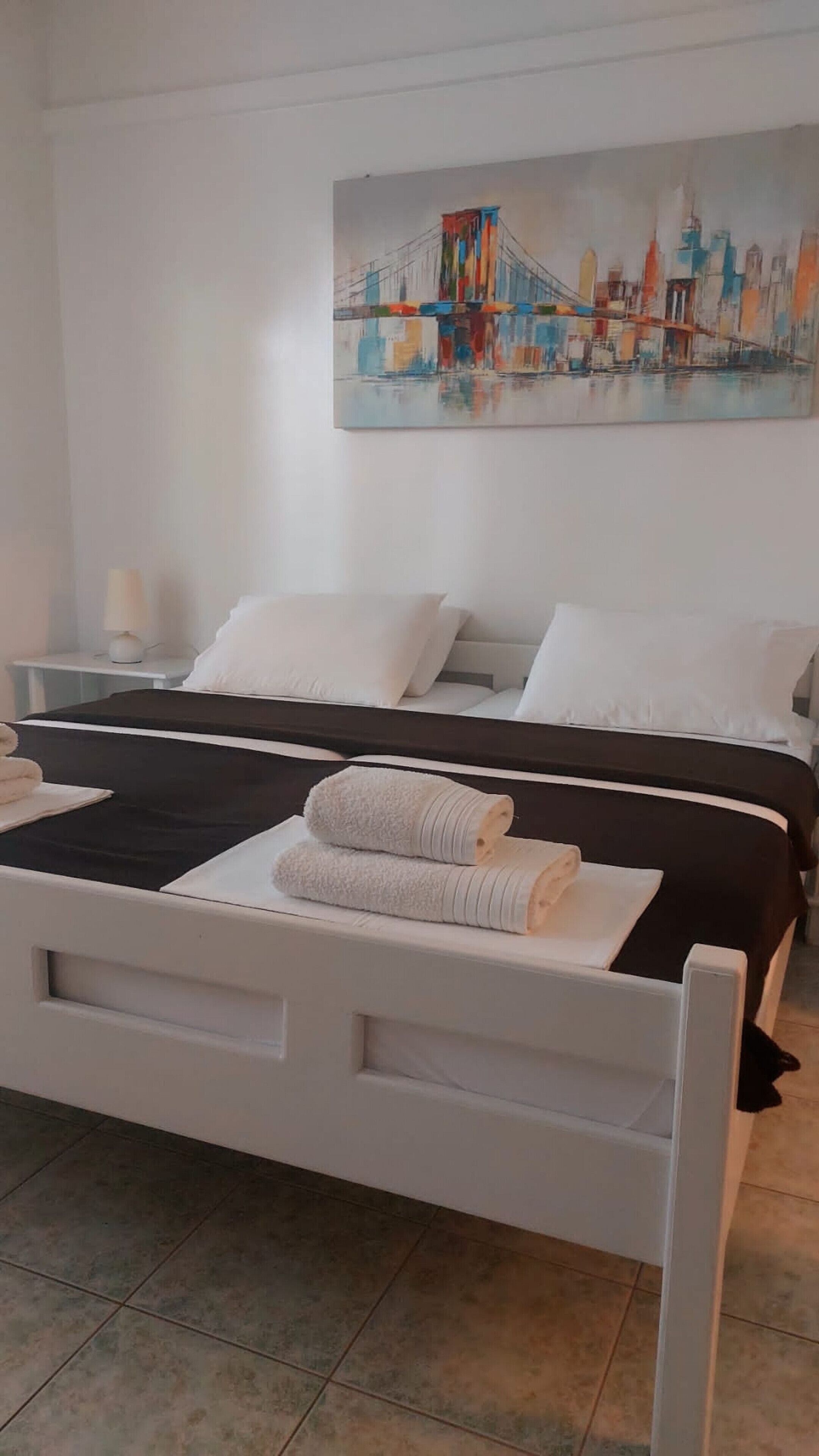 Una cama doble con ropa de cama blanca y negra, un cuadro de un paisaje urbano en la pared y una mesita de noche con una lámpara.