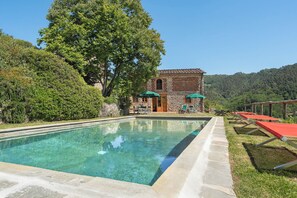 Ein Pool mit Steinrand, ein Steinhaus mit einer Tür und Blick auf einen bewaldeten Hügel.