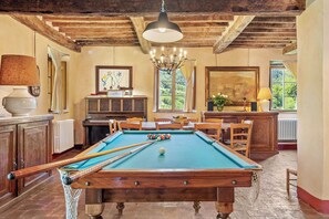 Une table de billard avec des billes, un piano et un lustre dans une pièce aux poutres apparentes et aux grandes fenêtres.