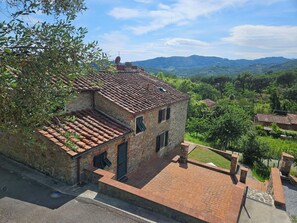 Un bâtiment en pierre avec un toit en tuiles, un petit porche et une vue sur un paysage verdoyant, avec des collines en arrière-plan.