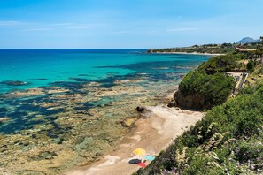 A coastal landscape with clear turquoise waters, rocky shorelines, and a path leading to a beach with an umbrella.