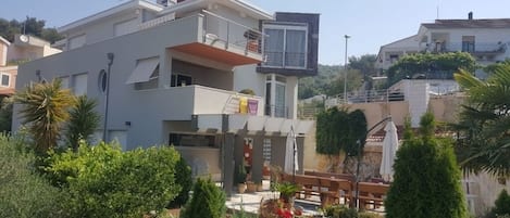 A modern two-story building with balconies, a garden area with potted plants, and a clear sky.