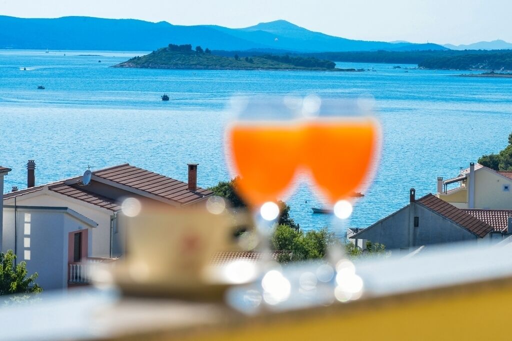 Un verre de jus d’orange au premier plan, avec en arrière-plan une vue sur une ville côtière et la mer.