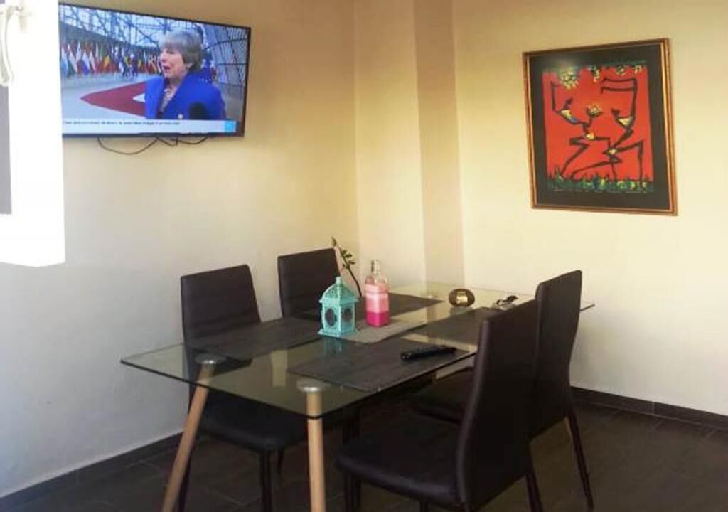 A dining area with a glass table, black chairs, a TV on the wall, and a framed abstract painting.