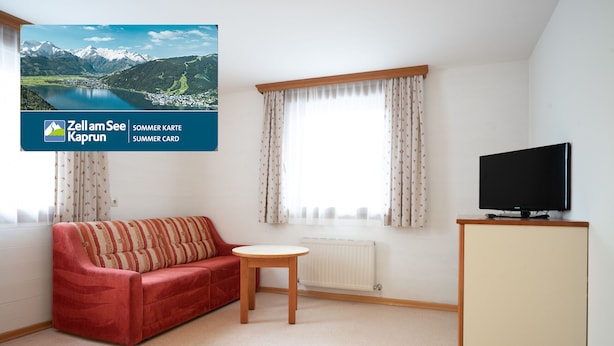 Een kamer met een rode bank, een klein rond tafeltje, een televisie op een kast en een raam met gordijnen.
