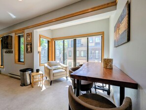 A room with a wooden dining table, a beige armchair, and a view of buildings through large windows. - Great Studio condo only steps from the Lifts! Hot Tubs and Fire Pit on site (Keystone)