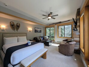A hotel room with a large bed, a bench, a sofa, a chair, a ceiling fan, and large windows with a view of trees. - Great Studio condo only steps from the Lifts! Hot Tubs and Fire Pit on site (Keystone)
