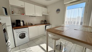 A modern kitchen with white cabinets, a built-in washing machine, a coffee maker, and a dining area with a wooden table and chairs. - Agréable T3 en Duplex dans résidence avec piscine (091) (Biscarrosse)