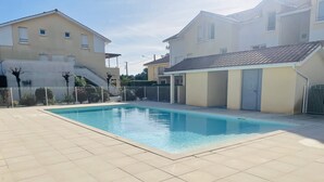 A rectangular swimming pool with tiled flooring, surrounded by a white fence and adjacent residential buildings. - Agréable T3 en Duplex dans résidence avec piscine (091) (Biscarrosse)