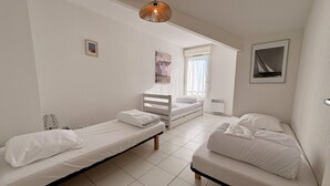 A small, clean room with two single beds, a wooden bench, a hanging light fixture, and a window with curtains. - Agréable T3 en Duplex dans résidence avec piscine (091) (Biscarrosse)