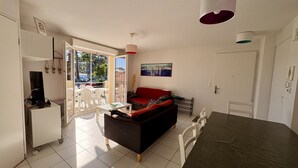 A modern living room with a sofa, dining table, and a TV. There is a balcony with a view of trees and parked cars. - Agréable T3 en Duplex dans résidence avec piscine (091) (Biscarrosse)