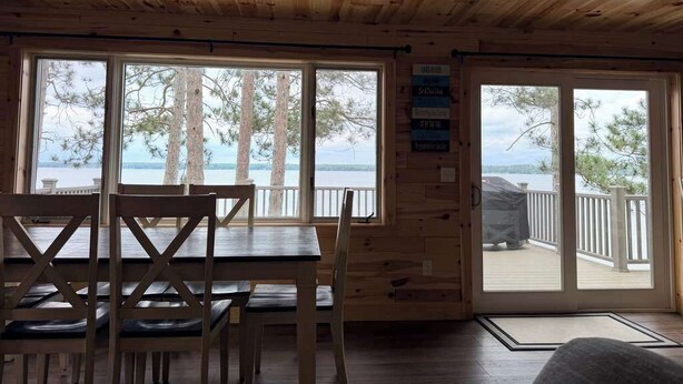 A wooden cabin interior with a dining table and chairs, large windows with a view of trees and water, and a deck area. - Veeder Lodge | West Twin Lake Retreat (Lewiston)