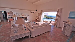 A spacious living room with a sofa, a coffee table, and a lamp. There is a bookshelf and a fireplace. A large window offers a view of the ocean. - Villa 10 people – Private infinity pool – Sea view – Wifi – Air conditioning – Sainte... (STE MAXIME)