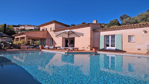 A swimming pool with lounge chairs and umbrellas, surrounded by buildings with terracotta roofs and light-colored walls. - Villa 10 people – Private infinity pool – Sea view – Wifi – Air conditioning – Sainte... (STE MAXIME)