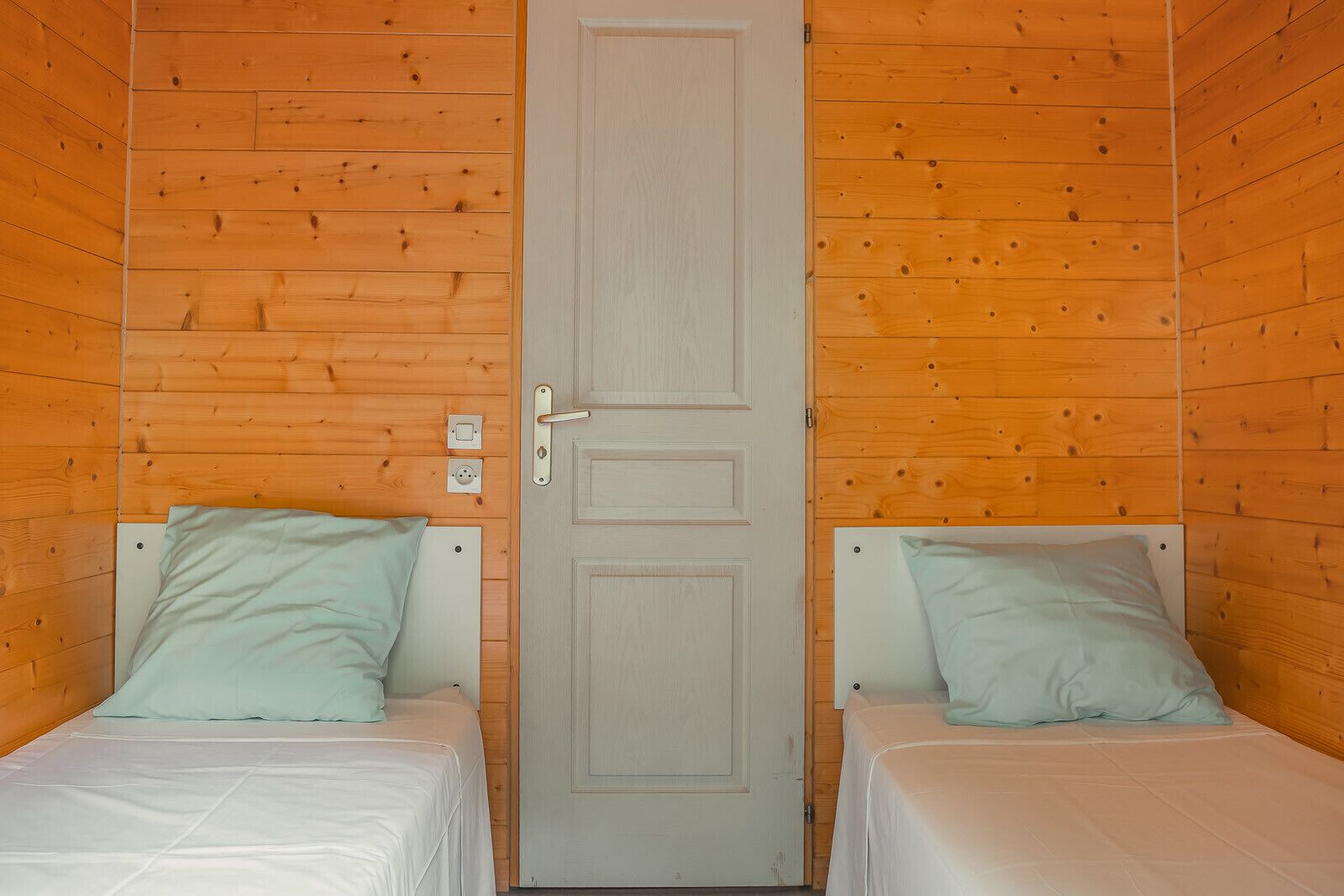 A room with two beds, each with a pillow and a white blanket. There is a door in the center with a handle.