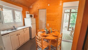 A wooden cabin with a kitchen featuring a sink, a microwave, and a refrigerator. There is a dining table with chairs and a window overlooking a deck.