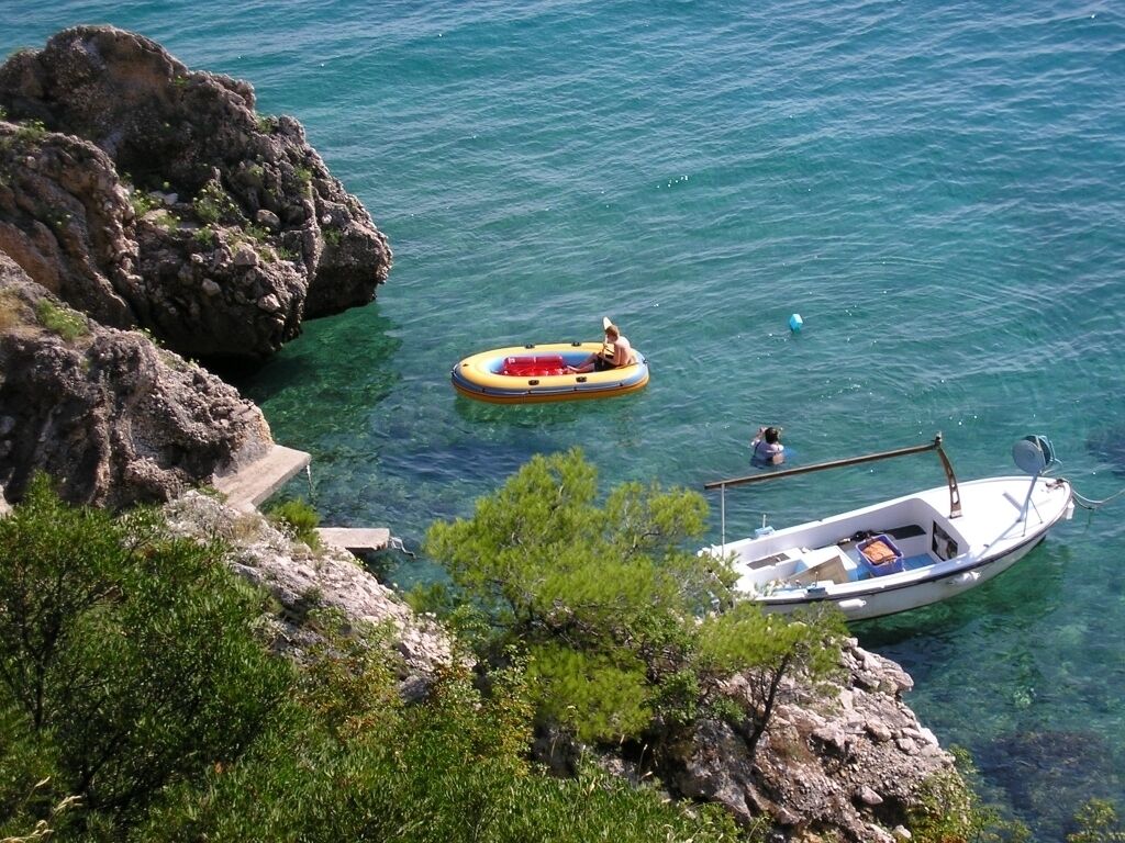 A rocky coastline with clear blue water, a small inflatable raft, and a white boat anchored nearby.