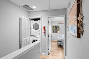 Un cuarto de lavandería con lavadoras y secadoras apiladas, un extintor y una puerta que da a un dormitorio con cama y ventana.