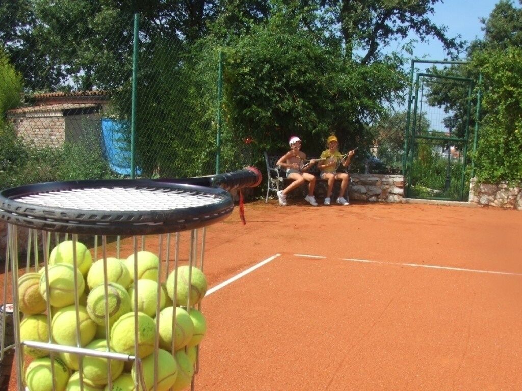 Ein Tennisplatz mit Netz, einem Ballnetz und zwei Personen, die im Hintergrund auf einer Bank sitzen.