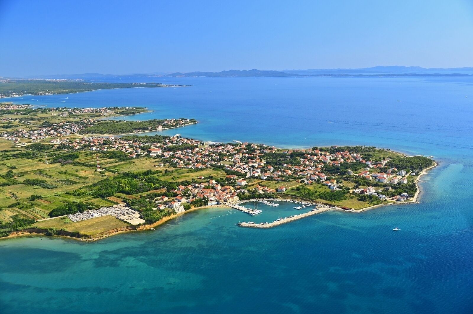 Vue aérienne d’une ville côtière avec un port de plaisance, des eaux d’un bleu limpide et une végétation luxuriante environnante.