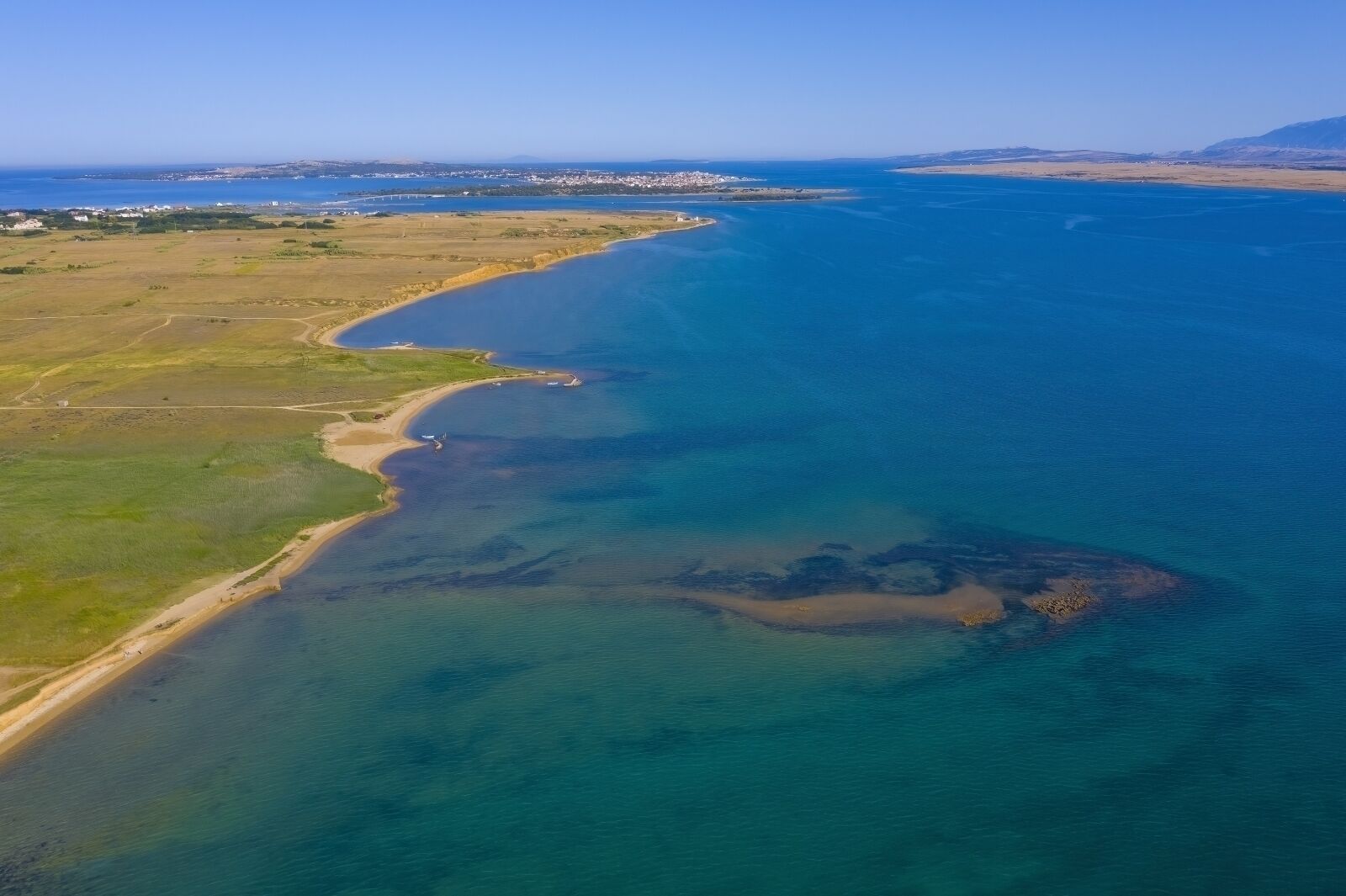 Vue aérienne d’une zone côtière avec une plage, des eaux d’un bleu limpide et des reliefs terrestres à l’horizon.