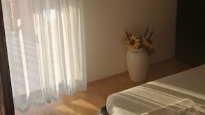 A bedroom with a bed, a window with sheer curtains, a mirror on the wall, and a vase with sunflowers.