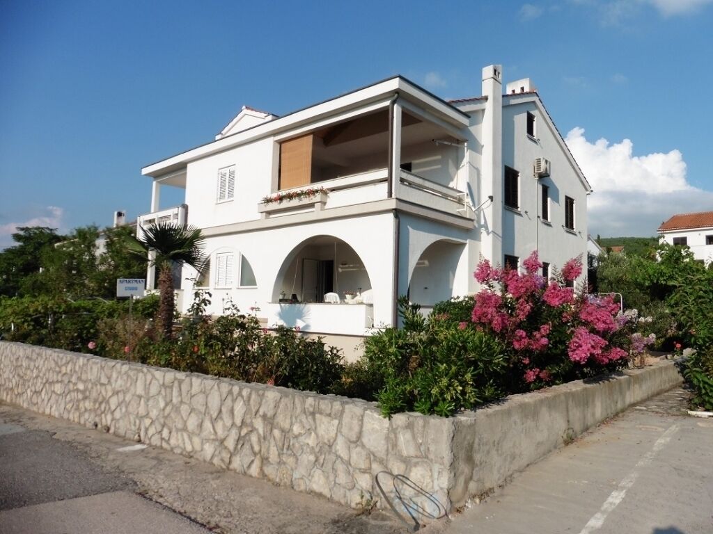 A two-story white building with a balcony, arched windows, and a stone wall in front.