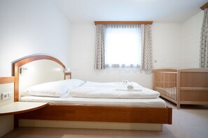 A bedroom with a wooden bed, a wooden headboard, a bedside table, a crib, and a window with curtains.