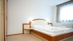 Ein ordentlich bezogenes Bett mit Holz-Kopfteil und Nachttisch, ein Heizkörper und ein Fenster mit Vorhängen.
