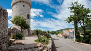 Una torre in pietra con base circolare e merlature, circondata da un percorso in pietra e vegetazione.